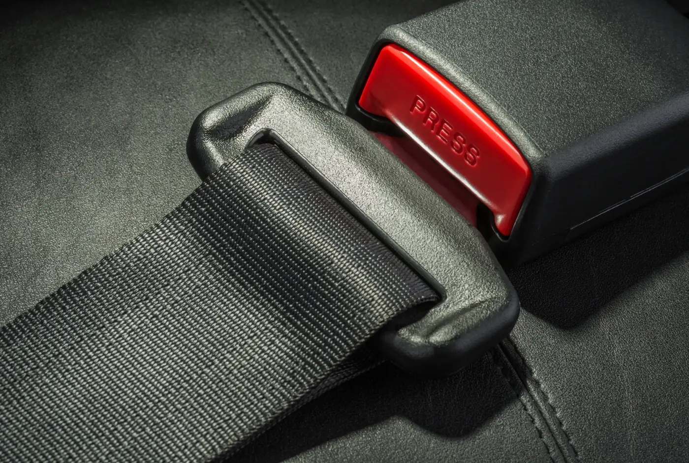 Seat belt tongue plate engaged in its buckle housing with the red release button visible, shown against dark upholstery